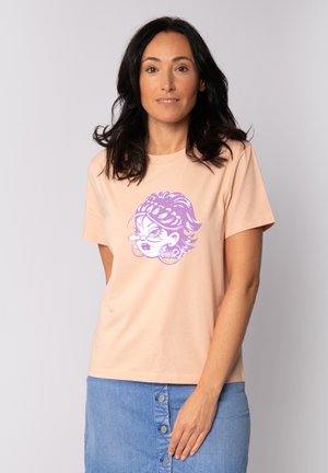 Femme aux cheveux foncés portant un t-shirt pêche avec un motif de visage de dessin animé violet et une jupe en denim bleue à boutons sur le devant, debout devant un fond clair.