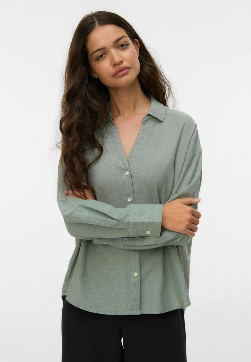 Femme aux longs cheveux bruns portant une chemise ample boutonnée vert sauge et un pantalon noir, debout les bras croisés sur un fond blanc.