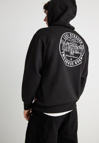 Άτομο που φοράει μαύρο φούτερ με κουκούλα με λευκό κυκλικό λογότυπο Levi Strauss Two Horse Brand στην πλάτη, στέκεται μπροστά σε απλό φόντο.