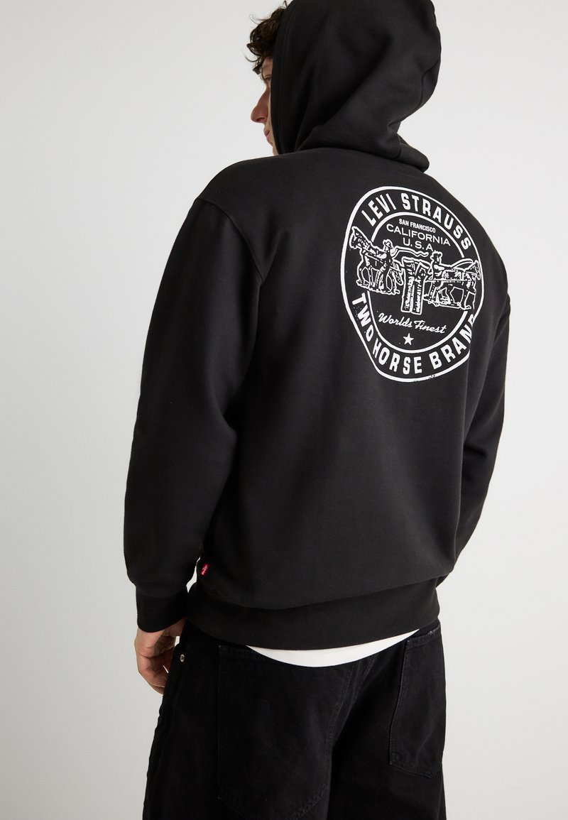 Άτομο που φοράει μαύρο φούτερ με κουκούλα με λευκό κυκλικό λογότυπο Levi Strauss Two Horse Brand στην πλάτη, στέκεται μπροστά σε απλό φόντο.