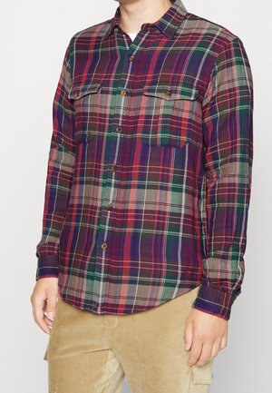 Homme portant une chemise à carreaux boutonnée à manches longues rouge, verte et bleue avec deux poches poitrine à l'avant et un pantalon en velours beige.