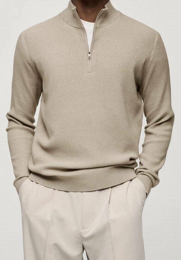 Homme portant un pull beige côtelé à demi-fermeture éclair sur une chemise blanche, associé à un pantalon plissé beige assorti, les mains dans les poches.