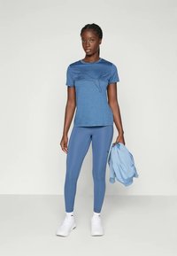 Μπλέ αθλητική μπλούζα με σκούρο μπλε γραφικό λογότυπο, σε συνδυασμό με μπλε κολάν και ανοιχτό μπλε μπουφάν, φορεμένα με λευκά αθλητικά παπούτσια.