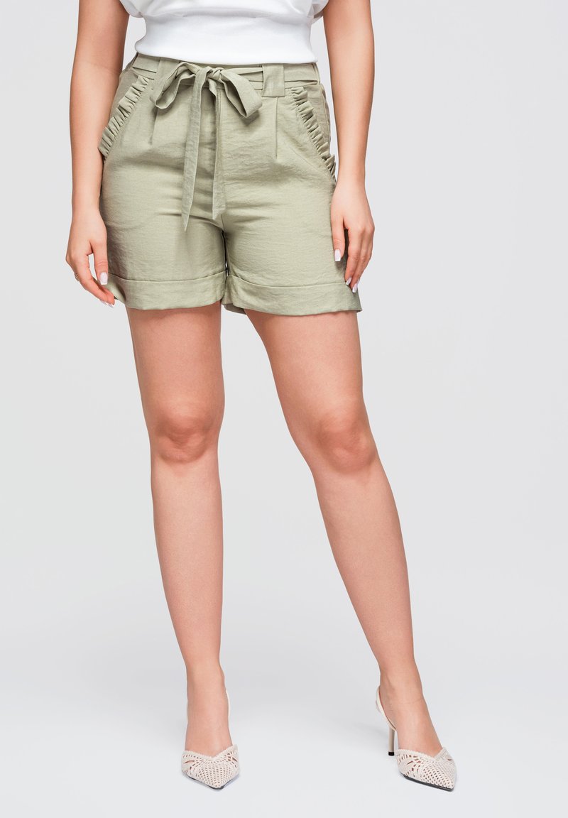 Femme portant un short taille haute vert clair avec une ceinture nouée et des poches froncées, associé à des talons pointus blancs en crochet.