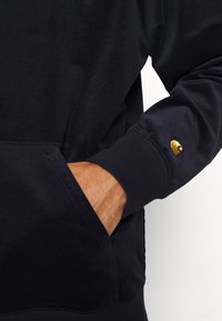 Navyblauer Pullover mit strukturierter Oberfläche, ausgestattet mit einer Fronttasche und einem goldenen Logo am Bündchen. Weiches Material und lässige Passform.