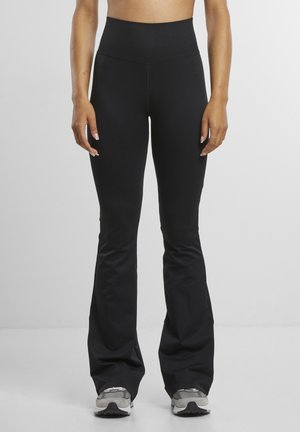 Femme portant des leggings noirs taille haute évasés et des baskets grises, debout devant un fond gris clair uni.