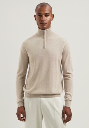 Beige ziptrui met lange mouwen, gemaakt van een zachte, gladde stof. Heeft een staande kraag en aansluitende manchetten, gecombineerd met lichte broek.