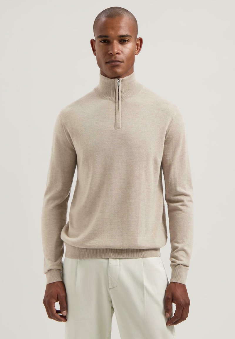 Beige svetr na zip s dlouhými rukávy, vyrobený z měkké, hladké látky. Má stojáček a přiléhavé manžety, v kombinaci se světlejšími kalhotami.