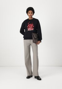 Sweatshirt preta com gráficos de corações vermelhos e texto branco, combinado com calças largas cinzentas e uma mala de mão castanha. Sapatos pretos completam o visual.