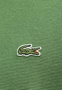Tissu tricoté vert avec un petit logo crocodile brodé, bouche rouge ouverte et contour blanc, centré sur le matériau.