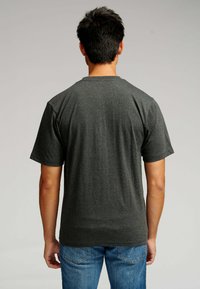 Mand med kort mørkt hår iført en ensfarvet mørkegrå T-shirt med korte ærmer og blå jeans, vendt væk mod en ensfarvet baggrund.