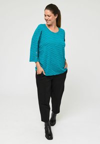 Teal teksturirana bluzica z valovitim vzorcem, ¾ rokavi z gumbom, v kombinaciji s črnimi ozkimi hlačami in gležnjarji. Minimalističen dizajn.