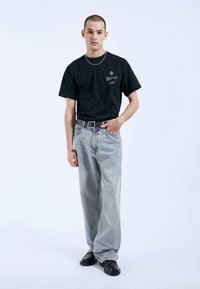 Camiseta de algodón negra con diseño gráfico, jeans de mezclilla azul claro, zapatos de cuero negros y collar de cadena plateada. Atuendo informal en tonos neutros.