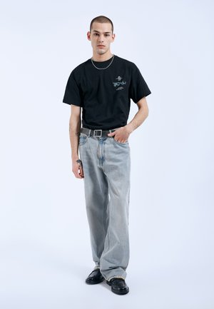 Ung mand iført sort T-shirt med grafik, lyseblå bukser med vide ben, sort bælte, sorte sko og et sølvkædehalskæde, stående med den ene hånd i lommen.