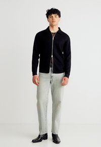 Νεαρός άνδρας στέκεται φορώντας μαύρο μπουφάν με φερμουάρ, ανοιχτό γκρι τζιν και μαύρα δερμάτινα παπούτσια μπροστά σε απλό ανοιχτό φόντο.