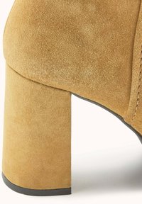 Chaussure à talon bloc en daim marron avec une surface texturée, une forme arrondie et une semelle en caoutchouc noire contrastante. Présente un détail de couture visible.