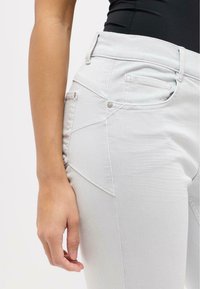 Light gray denim jeans with a fitted design, visible stitching details, and a side pocket. Material has a smooth texture, showing flexibility.