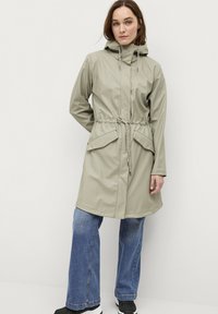Lichtgroene waterdichte jas met een capuchon, trekkoord in de taille en voorzakken. Gestyled met losse blauwe jeans en zwarte sneakers.
