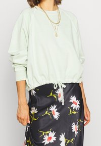 Ljust grön, kort sweatshirt med dragsko i nederkant, med ribbade muddarna och subtila logotyper. I kombination med en blommig svart kjol.
