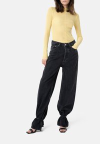 Langärmliges, eng anliegendes gelbes Oberteil kombiniert mit locker sitzenden, dunklen Jeans aus Denim, die eine hohe Taille und leicht ausgestellte Säume aufweisen. Schwarze, hochhackige Sandalen.