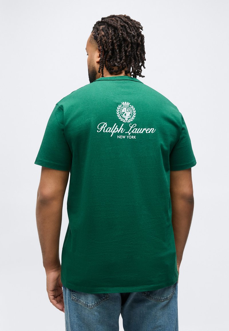 Άνδρας με κοντά dreadlocks γυρίζει την πλάτη, φοράει πράσινο t-shirt Ralph Lauren New York και μπλε τζιν, με φόντο απλό.