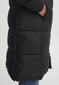 Svart pufferjakke med quiltet design, med lomme på siden og trykknapper. Myk, blankt stoff med en lett skinnende tekstur.