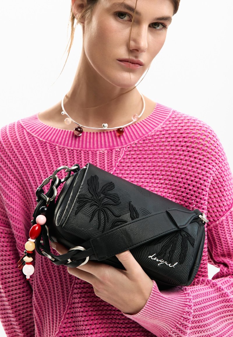 Femme portant un pull en maille rose et un collier de perles, tenant un sac à main en cuir noir avec un motif floral en relief et une sangle colorée en perles.