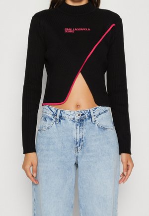 Mujer con un suéter negro corto con ribete rosa y logo, combinado con jeans azul claro de talle alto, mostrando el abdomen.