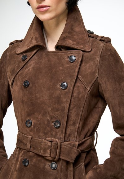 Close-up van een persoon die een bruine suède dubbelgeknoopte trenchcoat draagt met een brede kraag en een ceintuur in de taille.