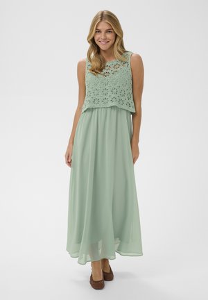 Lachende vrouw in een mouwloze maxi-jurk in saliegroen met een gebloemde gehaakte top en bruine gevlochten platte schoenen, staand op een witte achtergrond.