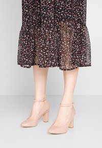 Beiga lacklädershöga klackskor med rund tå och ankelrem, med en fyrkantig blockklack och en slät yta.