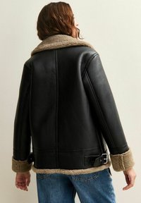 Persona con capelli castani indossa una giacca di pelle nera con colletto e polsini in shearling beige, abbinata a jeans blu, voltata di spalle rispetto alla fotocamera.
