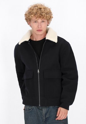 Black zip-up jacket with a cream shearling collar, two front pockets, and a soft texture, paired with a black t-shirt and blue denim jeans.