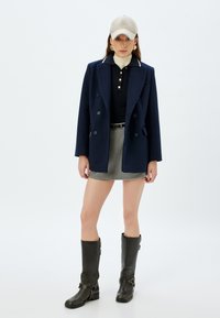 Cappotto doppiopetto blu navy con colletto bianco, maglietta nera, gonna mini in panno scozzese, stivali neri in pelle alti e cappello beige.