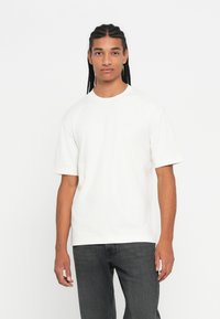 Νεαρός άνδρας με μακριά πλεγμένα μαλλιά, φορώντας ένα απλό λευκό T-shirt με κοντά μανίκια και σκούρα γκρι τζιν, που στέκεται μπροστά σε λευκό φόντο.