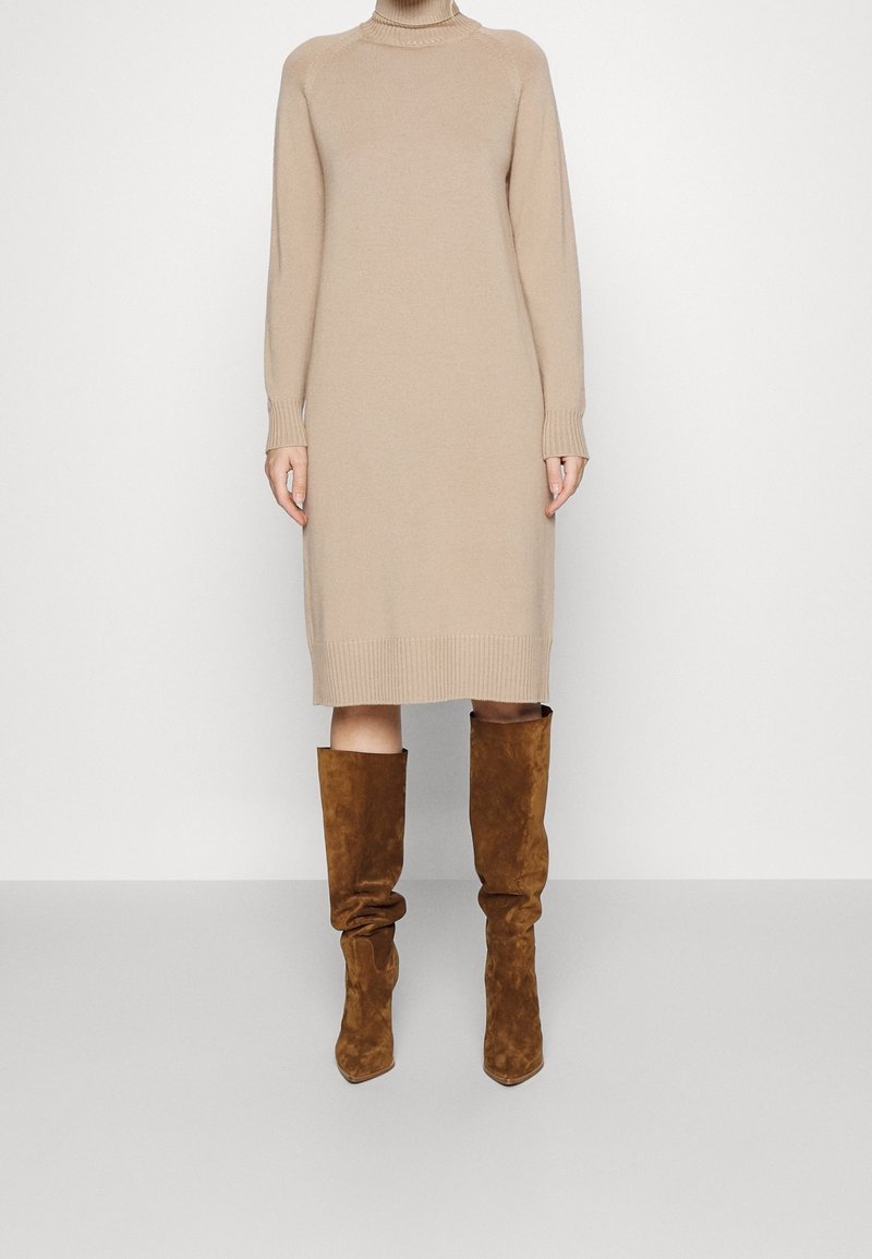 Robe en maille beige à manches longues avec col roulé, associée à des bottes en daim marron décontractées montant jusqu'aux genoux, sur un fond uni.