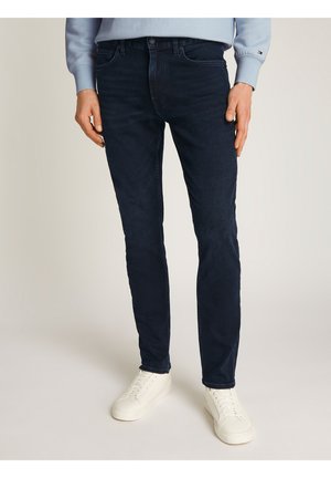 Homme portant un jean slim foncé, un sweat-shirt bleu clair et des baskets blanches, debout sur un fond clair.