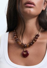 Collana di perle marroni con un pendente circolare rosso, caratterizzata da una texture naturale e un design a strati. Indossata sopra una maglietta bianca.