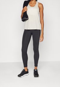 Top deportivo de color beige claro con un pequeño logo, combinado con leggings negros ajustados y zapatillas deportivas negras. Toalla colgada del hombro.