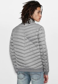 Grå pufferjakke med chevron-quiltet design og ribbede manchetter. Har en mærket elastisk kant og en glat, shiny tekstur.