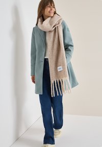 Teal kappa med en strukturerad beige scarf som har fransar och ett logomärke, i kombination med mörkblå byxor och ljusa sneakers.
