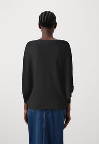 Haut en tricot noir texturé avec un col large et une coupe ample, associé à une jupe en denim bleu, vu de dos.