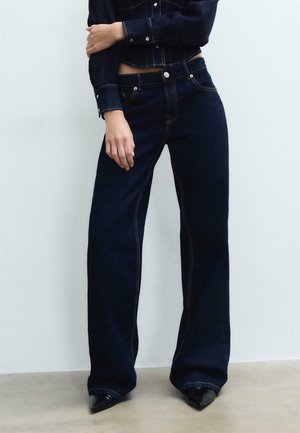 Personne portant un jean large bleu foncé et une veste en jean courte assortie, debout devant un fond uni gris clair.