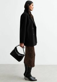 Manteau noir à double boutonnage, jupe midi imprimé animal, bottes noires jusqu'aux genoux et un petit sac à main noir au design arrondi et à poignée courte.
