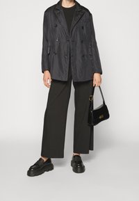 Veste noire légère à double boutonnage avec une finition satinée, associée à un pantalon large noir, des chaussures noires robustes et un sac à main noir.