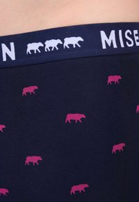 Boxers pour hommes en marine avec une ceinture douce. Présentent des silhouettes de vaches roses répétées sur le tissu. Texture lisse, design décontracté.