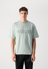 Mlad moški s kratkimi rjavimi lasmi, oblečen v svetlo sivo preveliko majico z logotipom "HUGO" in črnimi hlačami, stoji pred enobarvno belo ozadjem.