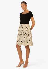 Abito nero off-the-shoulder con corpino aderente e gonna crema al ginocchio con ricami floreali e orlo scallop. Sandali con tacco trasparente.
