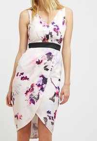 Abito avvolgente floreale in crema chiaro con motivi acquerello viola e rosa, caratterizzato da una cintura nera e una scollatura a V. Finitura in tessuto liscia.