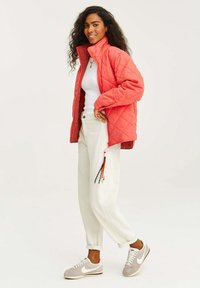 Kurtka z pikowanego koralowego materiału z wysokim kołnierzem, dopasowana biała bluzka, beżowe spodnie z podwiniętymi nogawkami i ozdobnymi frędzlami oraz szare trampki.